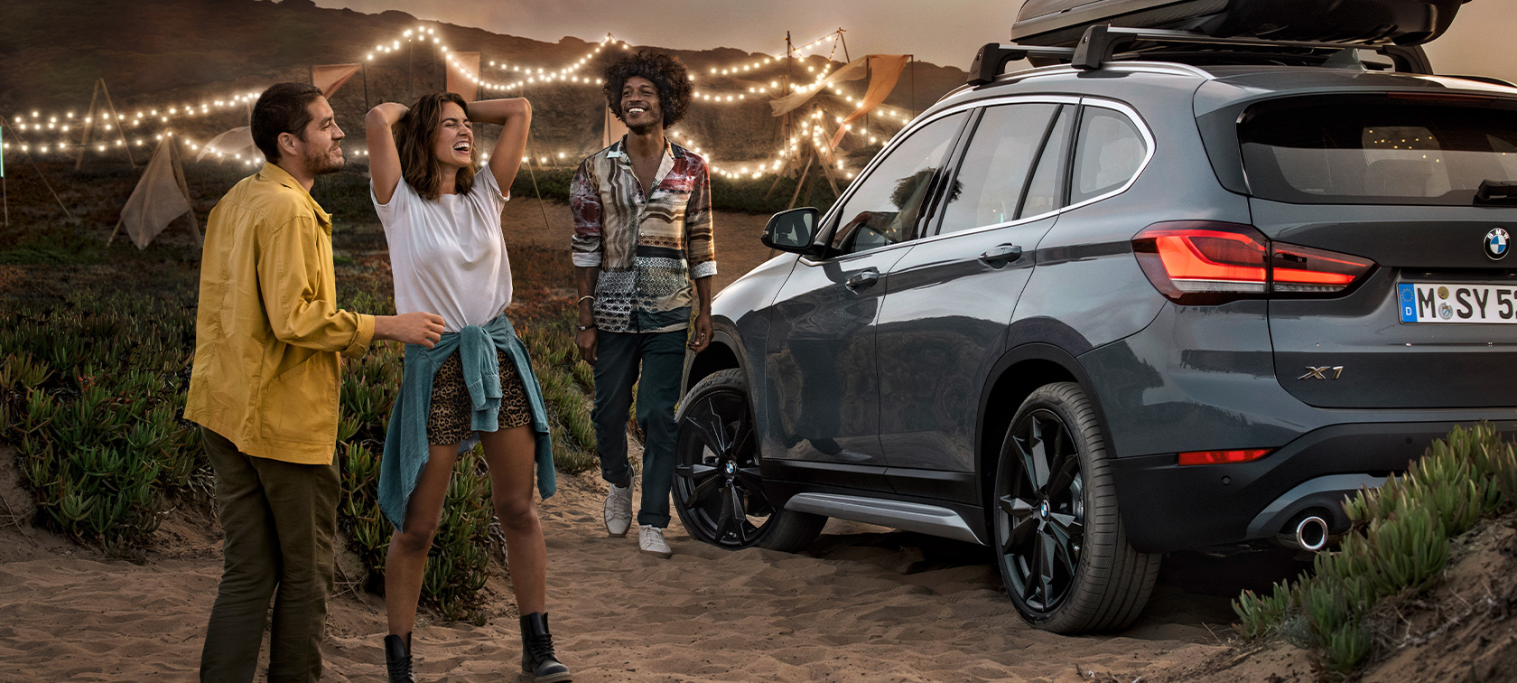 Oslavujúce trio v BMW X1 s BMW letnými ponukami Šťastné trio v BMW X1 Mineral Grey vybavenom BMW letnými ponukami trojštvrťový zadný pohľad na pláži BMW vybavenie