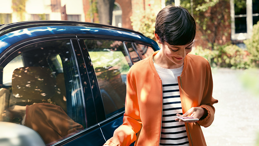 BMW letné ponuky BMW Teleservices BMW letné ponuky BMW Teleservices žena stojaca pred BMW, ktorá používa smartfón