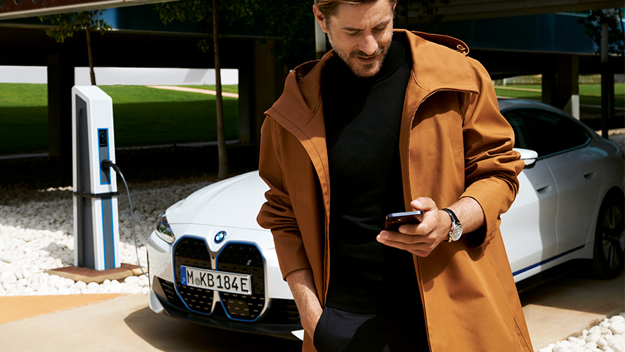Trvanie a zmluvné obdobia balíkov BMW Service Inclusive pre elektrické vozidlá Muž s balíkom BMW Service Inclusive pozerá na smartfón pred BMW i4 na nabíjacej stanici