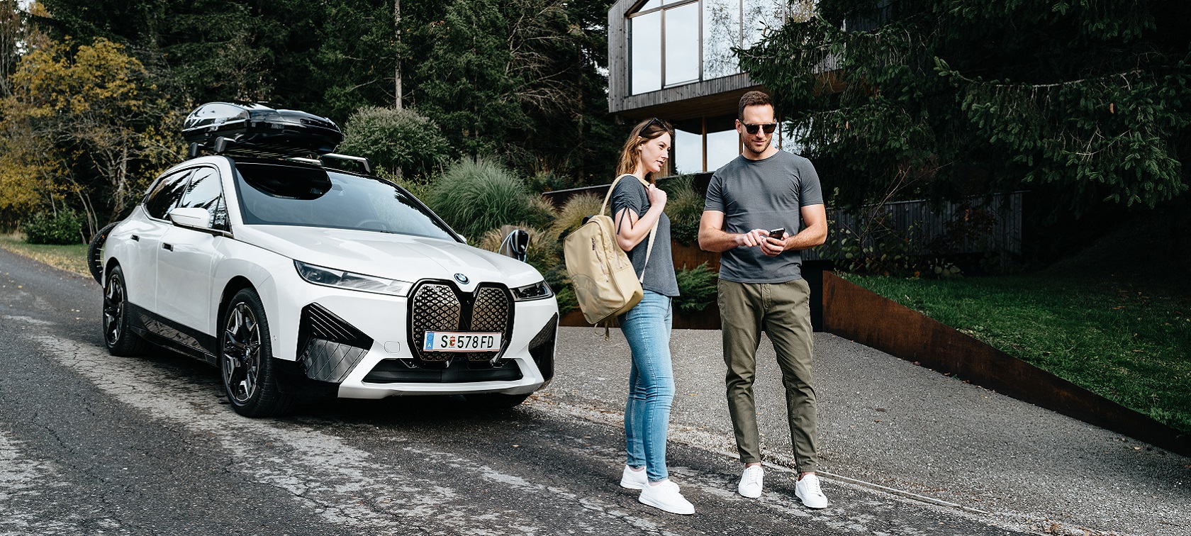 Muž s telefónom a žena s batohom stoja pred BMW iX. Muž s telefónom a žena s batohom stoja pred BMW iX.