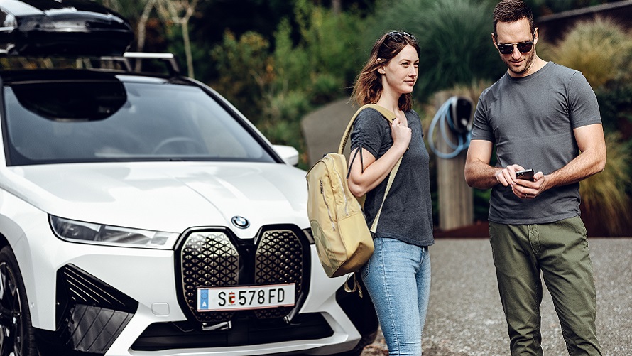 Muž s telefónom a žena s batohom stoja pred BMW iX. Muž s telefónom a žena s batohom stoja pred BMW iX.