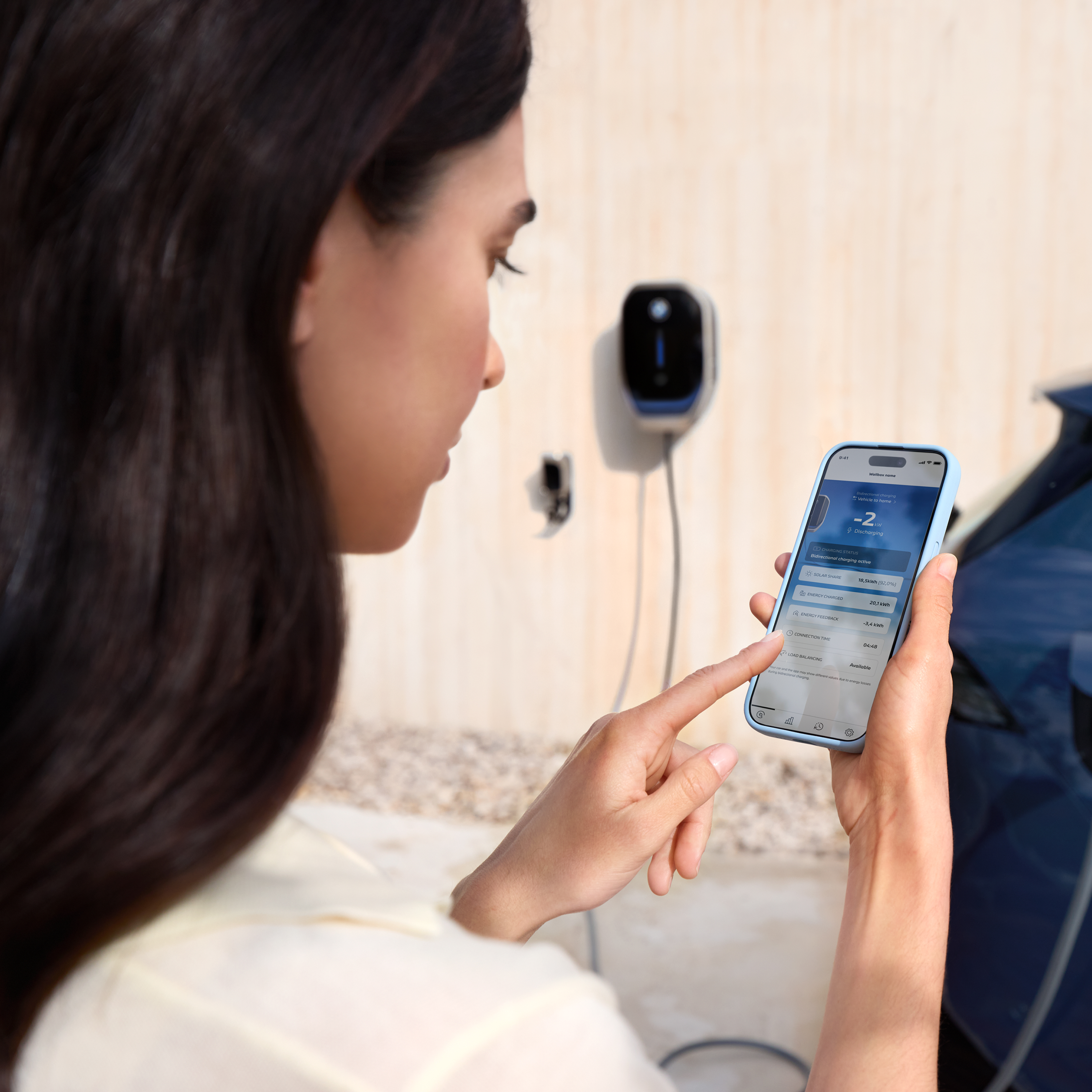 Žena sa pozerá na svoj smartfón, na pozadí je viditeľný wallbox